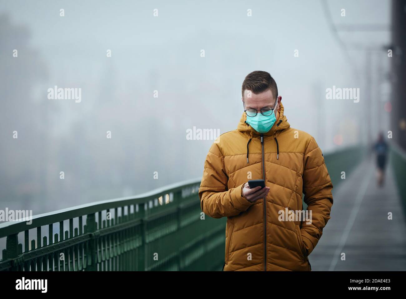 Einsamer Mann mit Gesichtsmaske mit Telefon während des Spaziergangs auf Brücke gegen die Stadt in geheimnisvollen Nebel. Düsteres Wetter in Prag, Tschechische Republik. Stockfoto