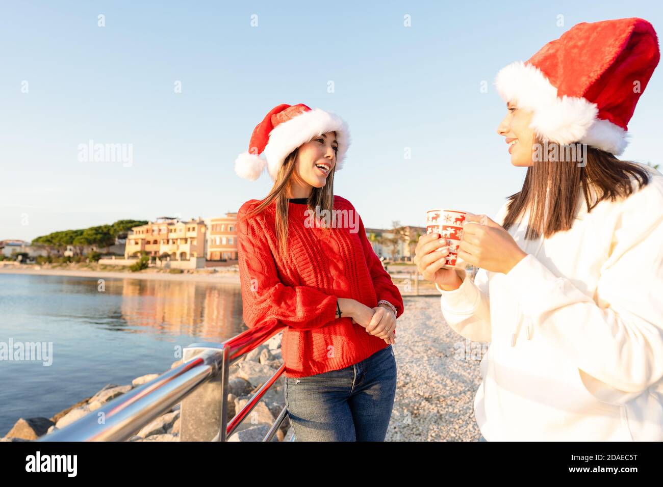 Schöne junge kaukasische Frauen stehen im Freien und sprechen mit einem Weihnachtsfest Tasse in den Händen tragen Weihnachtsmütze - Sonnenuntergang Meer Winter Urlaub für zwei Frauen f Stockfoto
