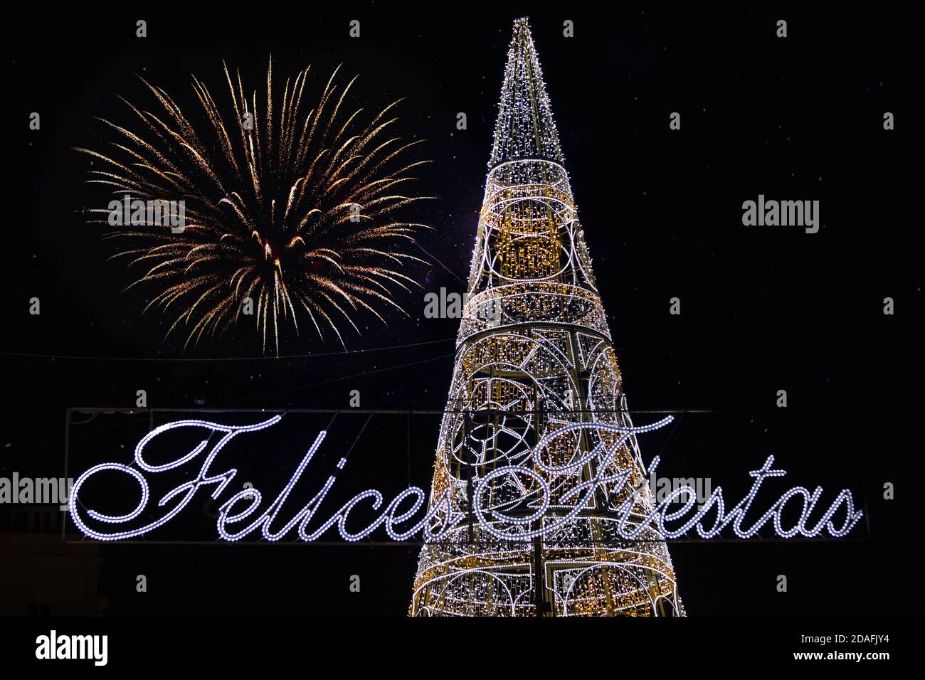 Weihnachtsbaum beleuchtet neben dem Text Frohe Feiertage in Spanisch geschrieben und Feuerwerk am Himmel. Stockfoto