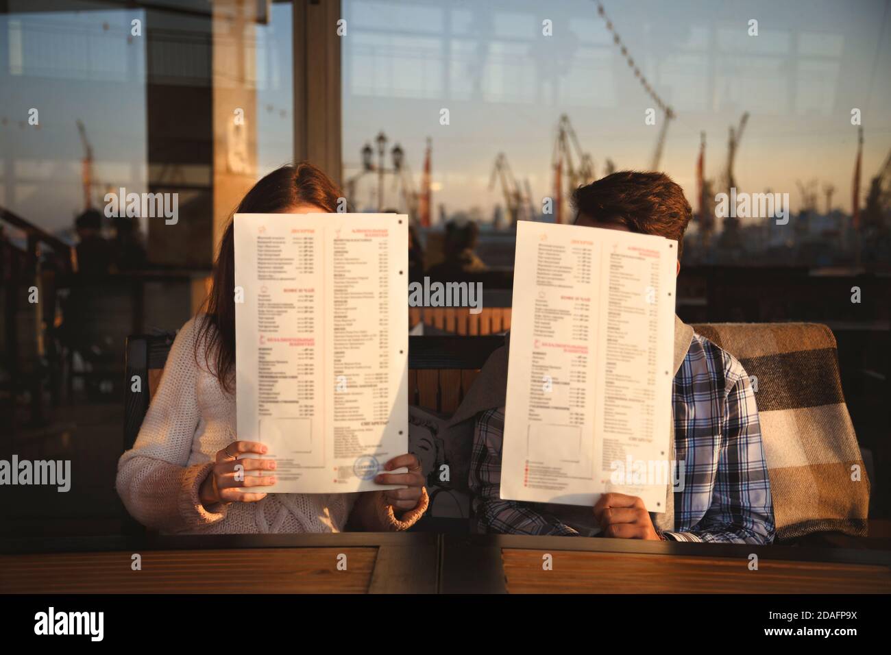 Junge lustige glücklich liebende Paar sitzt am Meer Hafen Open-Air-Café und täuschen ihre Gesichter mit Menüs zu schließen. Beginn der Liebesgeschichte. Beziehung Stockfoto