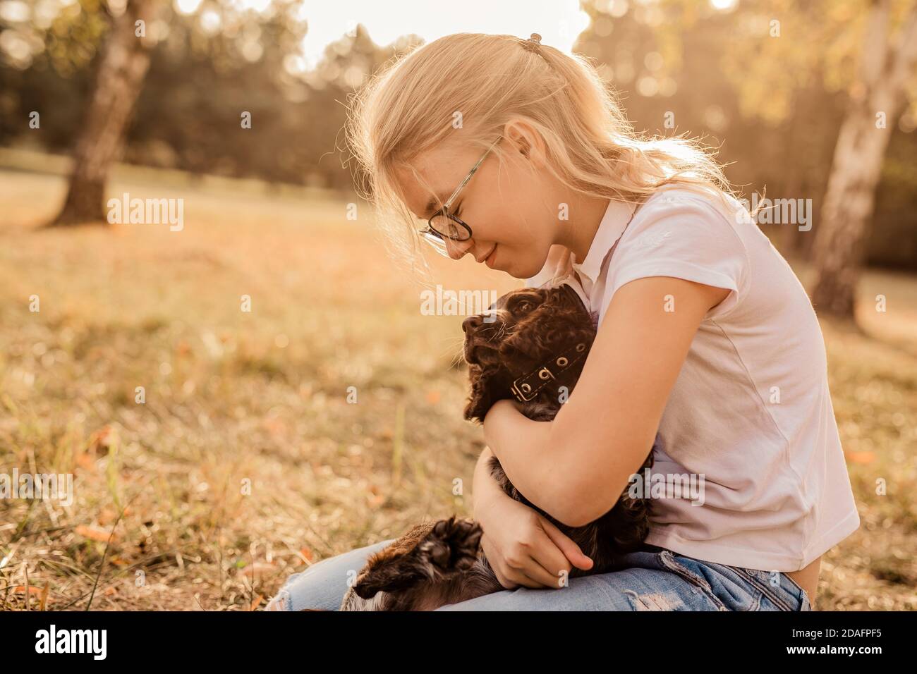 12 Jahre alt blonde Mädchen mit großen Brillen lachen und Spielen mit kleinen Welpen Spaniel im warmen Herbst Park Oder Wald Stockfoto