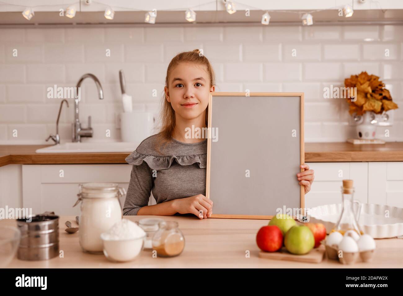 Teenager-Mädchen in einem grauen Kleid wird kochen Ein traditioneller Apfelkuchen in der neuen Küche und hält Ein leeres Holzregal mit grauem Hintergrund mit einem Stockfoto