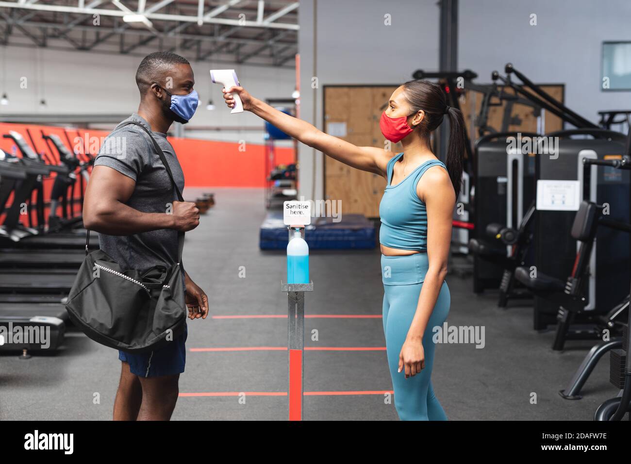 Fit kaukasische Frau trägt Gesichtsmaske Überprüfung der Temperatur der Passform afroamerikanischer Mann mit Sporttasche Stockfoto