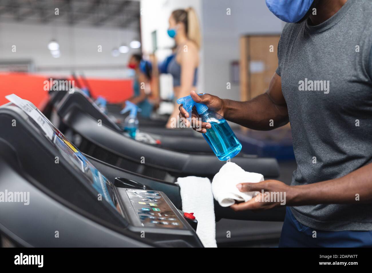 Mittelteil des fitfähigen afroamerikanischen Mannes mit Gesichtsmaske Desinfizieren des Cardio-Geräts vor der Arbeit o Stockfoto