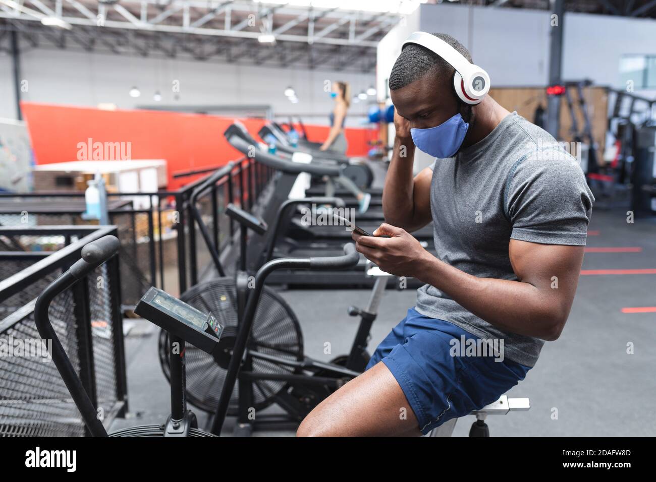 Fit afroamerikanischen Mann trägt Gesichtsmaske und Kopfhörer mit Smartphone, während Sie auf der Stationa sitzen Stockfoto