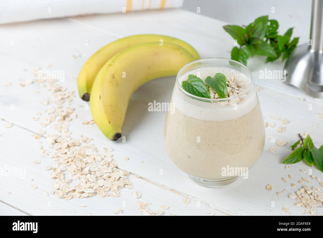 Bananenmilchshake mit Hafermilch. Auf dem rustikalen weißen Holztisch. Im Cocktail sind ganze Haferkörner und Minze für Frische. Morgendlicher Smoothie. Stockfoto
