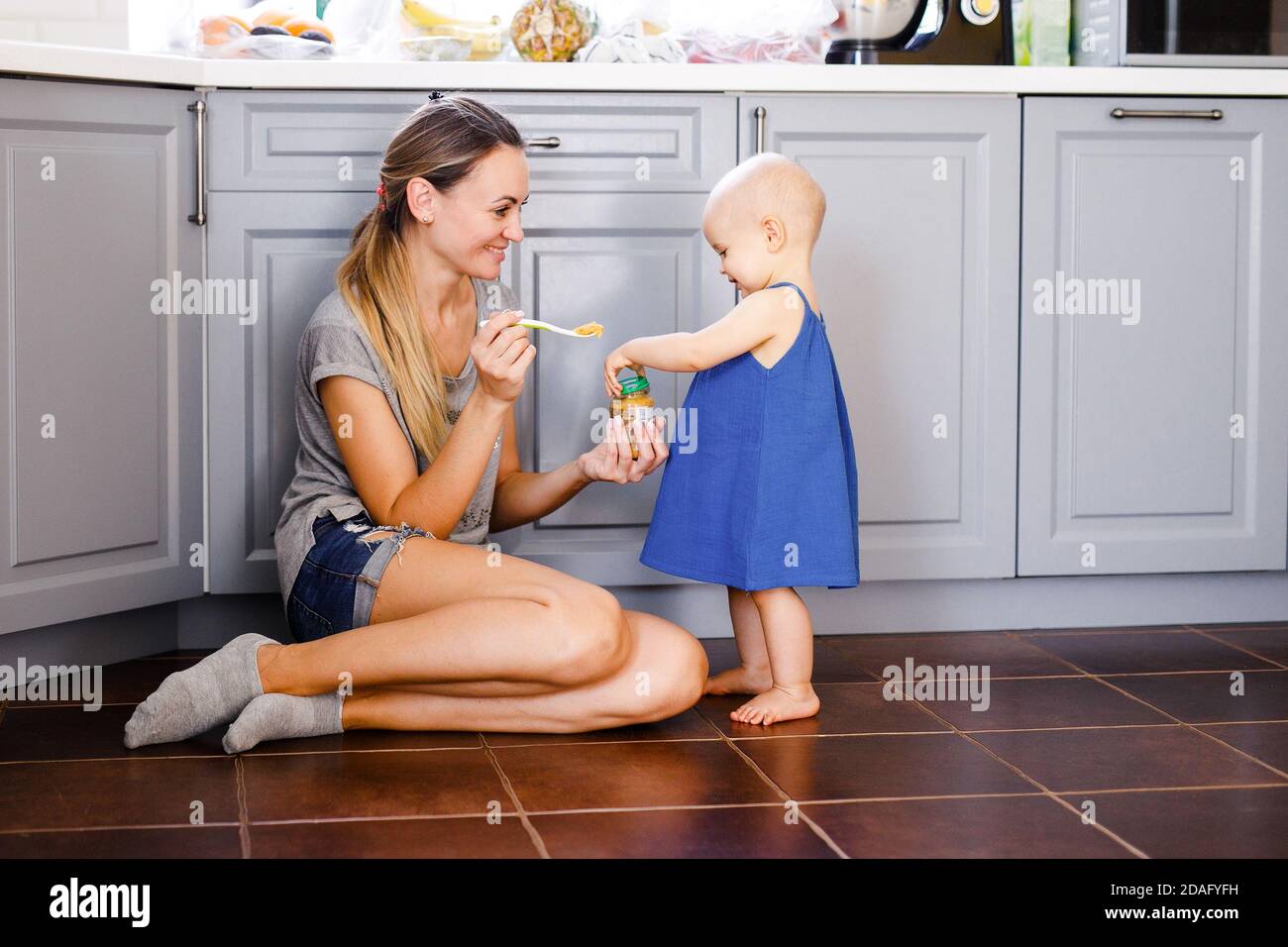 Eine junge Mutter sitzt auf dem Boden in der Küche und füttert ihre einjährige Tochter mit einem Löffel. Stockfoto