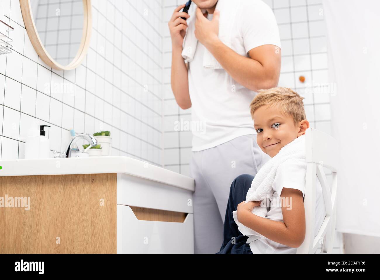 Positiver Junge, der die Kamera anschaut, während er auf einem Stuhl in der Nähe sitzt Mann rasieren mit elektrischen Rasierer im Badezimmer Stockfoto