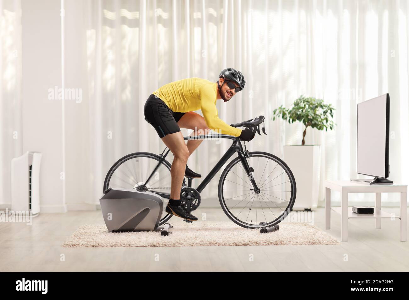 Ganzkörperaufnahme eines jungen Mannes, der mit einem Fahrrad Trainer Fahrrad zu Hause und Blick auf die Kamera Stockfoto