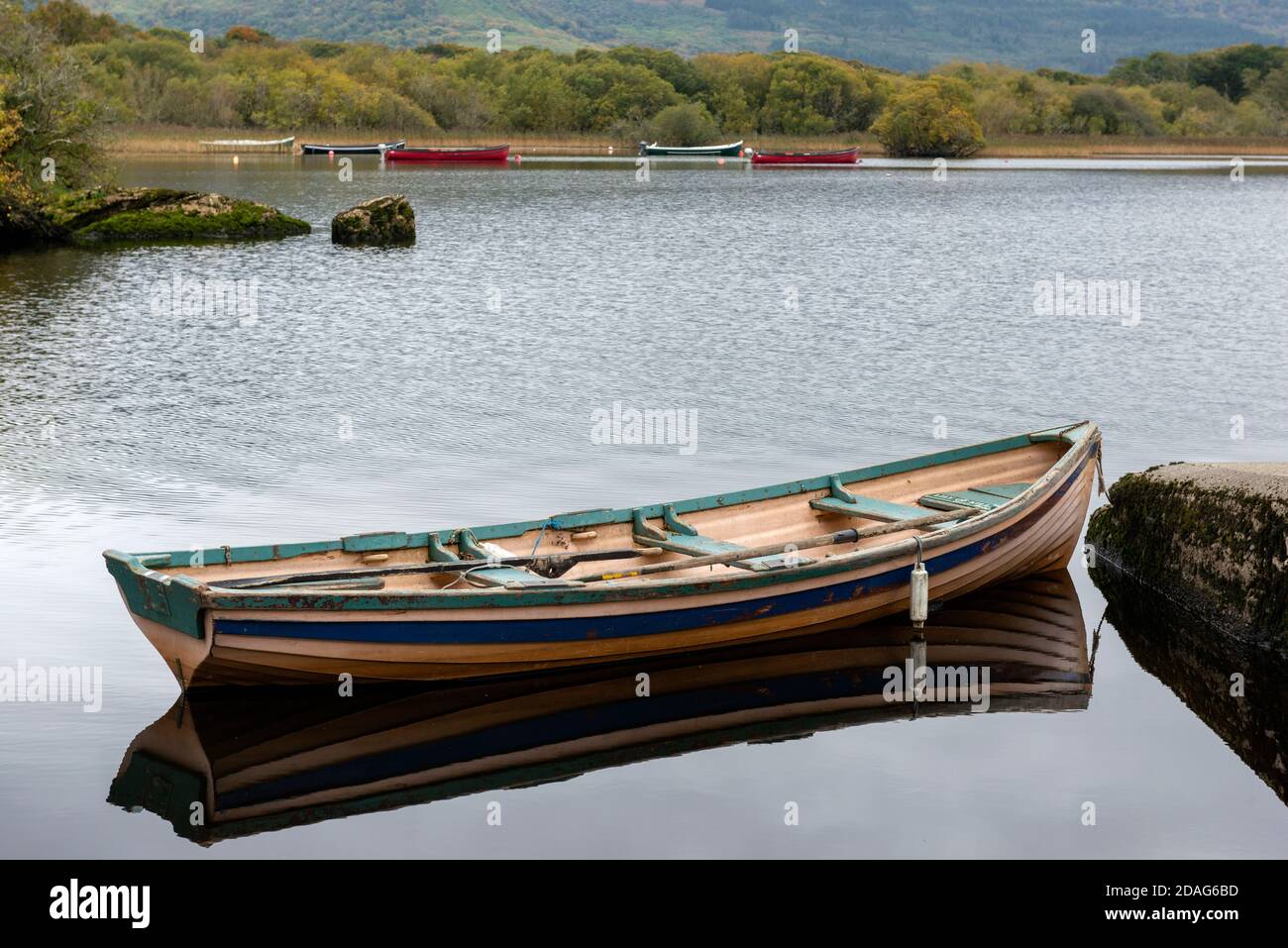 Ein kleines Ruderboot oder Klinkerboot auf einem ruhigen See gegen verankerte Fischerboote im Lough Leane im Killarney National Park, County Kerry, Irland Stockfoto