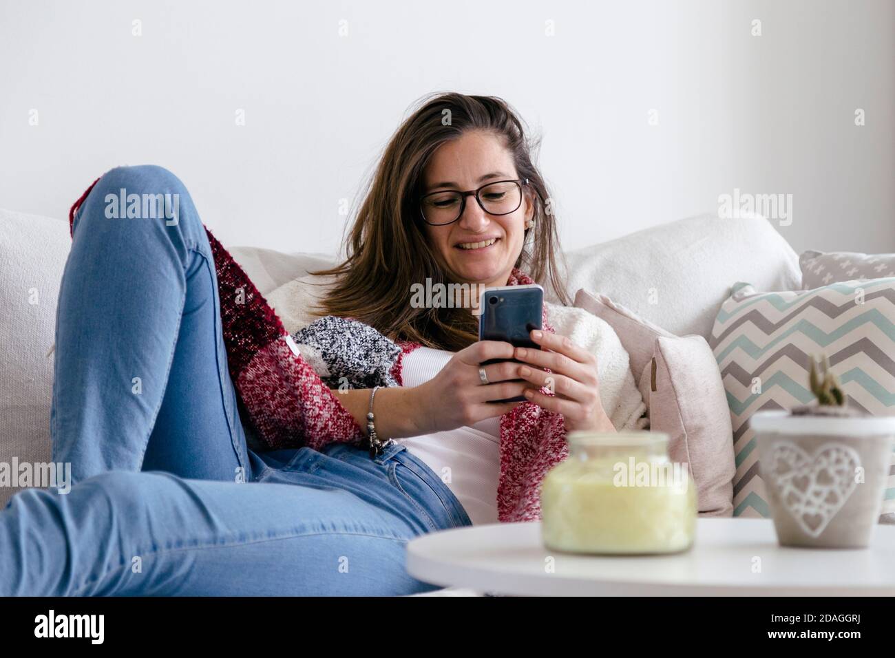 Lächelnde junge Frau zu Hause entspannt auf dem Sofa mit einem Handy in den Händen. Stockfoto