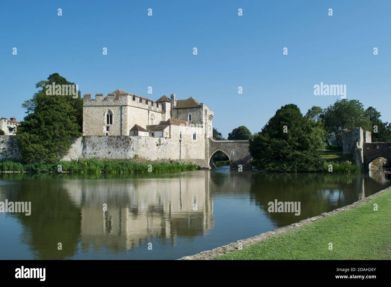 Eine europäische Steinburg (befestigtes Herrenhaus) auf einem See oder Fluss (Graben), reflektiert in der ruhigen Wasser durch helles Sonnenlicht. Bäume, Grasbänke und Stein Stockfoto