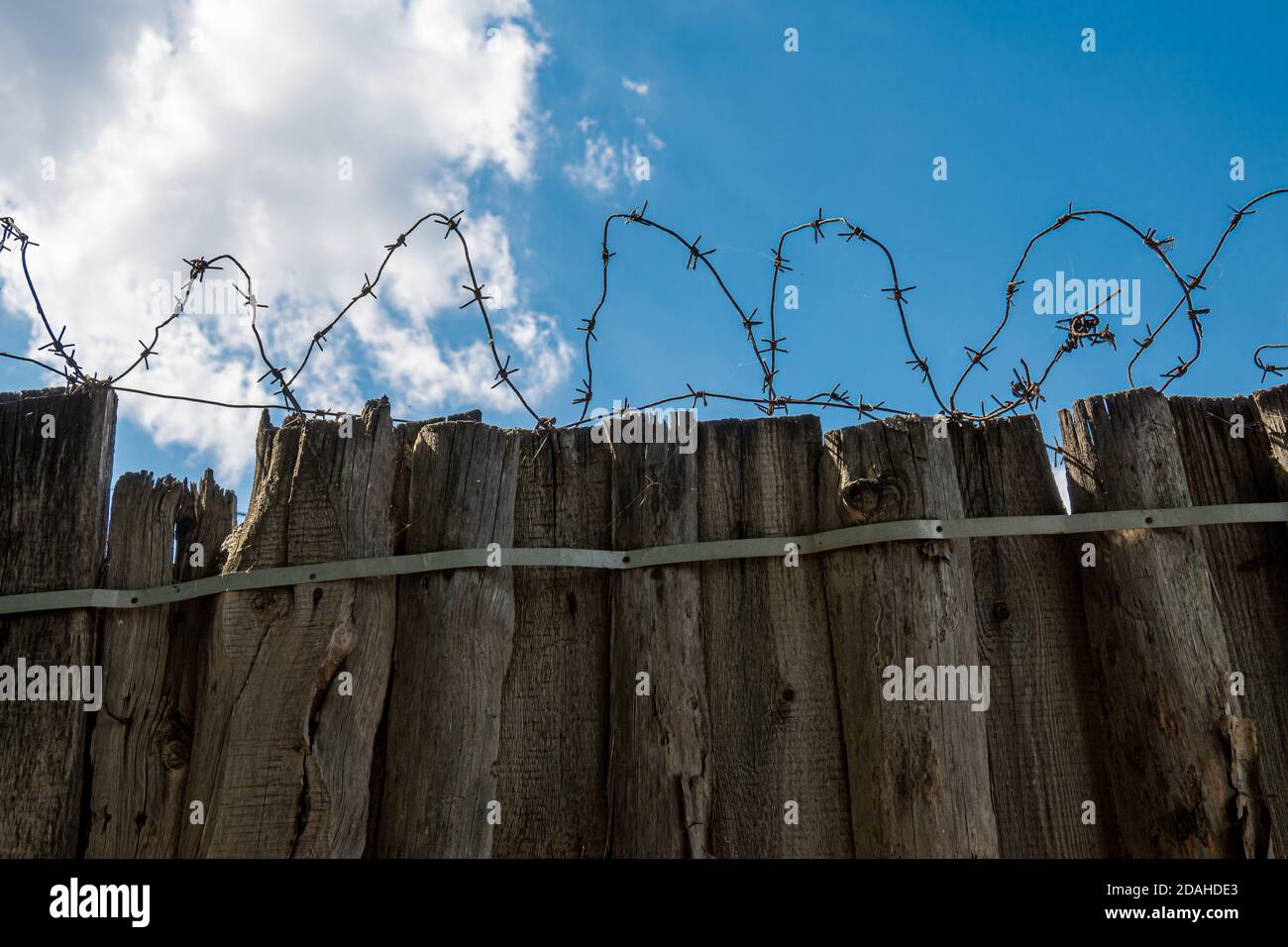 Die Silhouette von Windungen aus altem rostigen Stacheldraht auf einem alten abgenutzten Holzzaun gegen einen strahlend blauen Himmel und Wolken. Speicherplatz kopieren. Stockfoto