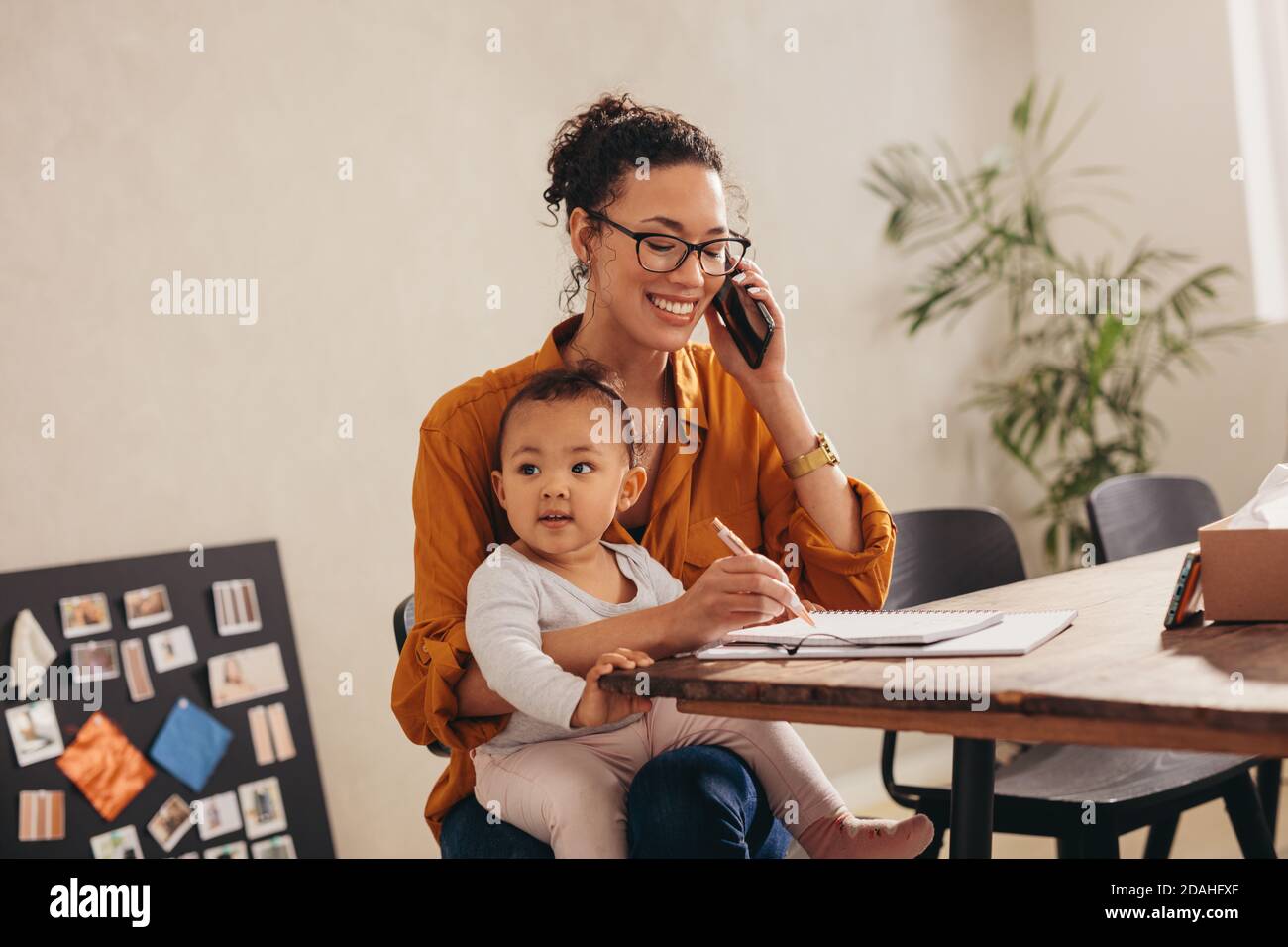 Geschäftsfrau telefoniert mit ihrem Baby, das zu Hause auf ihrem Schoß sitzt. Arbeitende Mutter macht Notizen, während sie auf dem Mobiltelefon spricht. Stockfoto