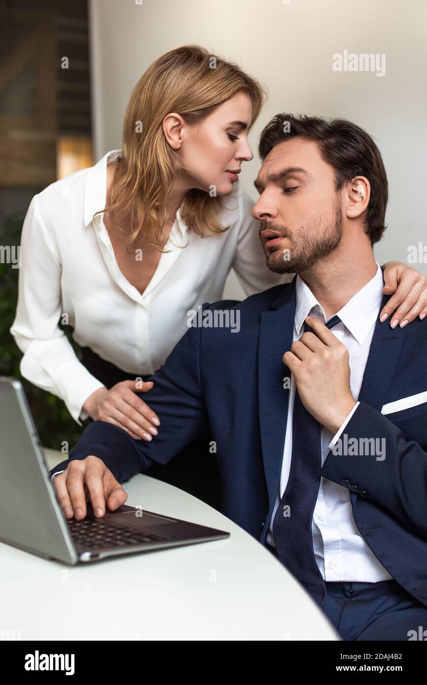 Leidenschaftliches Paar flirtet im Büro. Geschäftsmann sieht auf Frau, die ihn von hinten berührt, während sie in der Nähe des Arbeitsplatzes steht. Verführerische Konzept Stockfoto