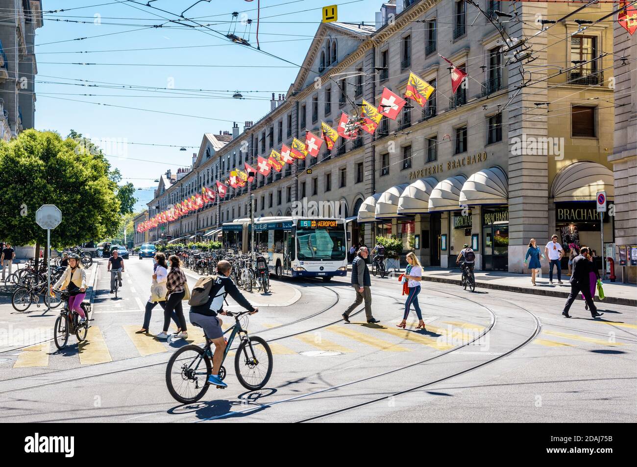 Fußgänger und Radfahrer überqueren die Rue de la Corraterie in Genf, deren Gebäude mit Fahnen geschmückt sind, während ein Bus an einer roten Ampel wartet. Stockfoto