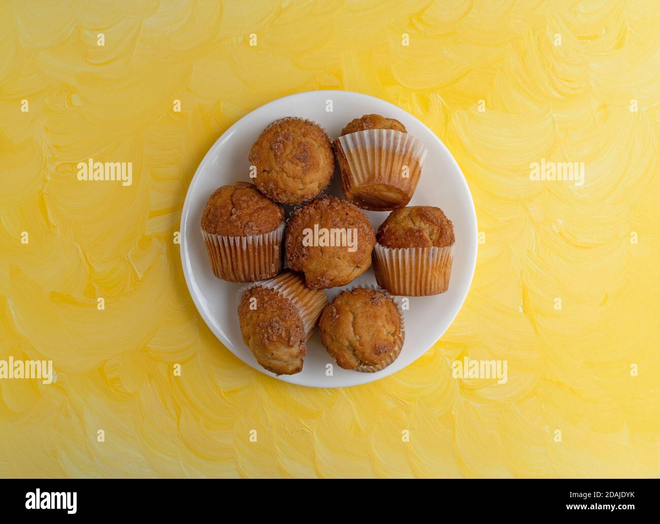 Draufsicht auf mehrere Bananenstreusel-Muffins auf einer weißen Platte auf einer gelb lackierten Tischplatte. Stockfoto