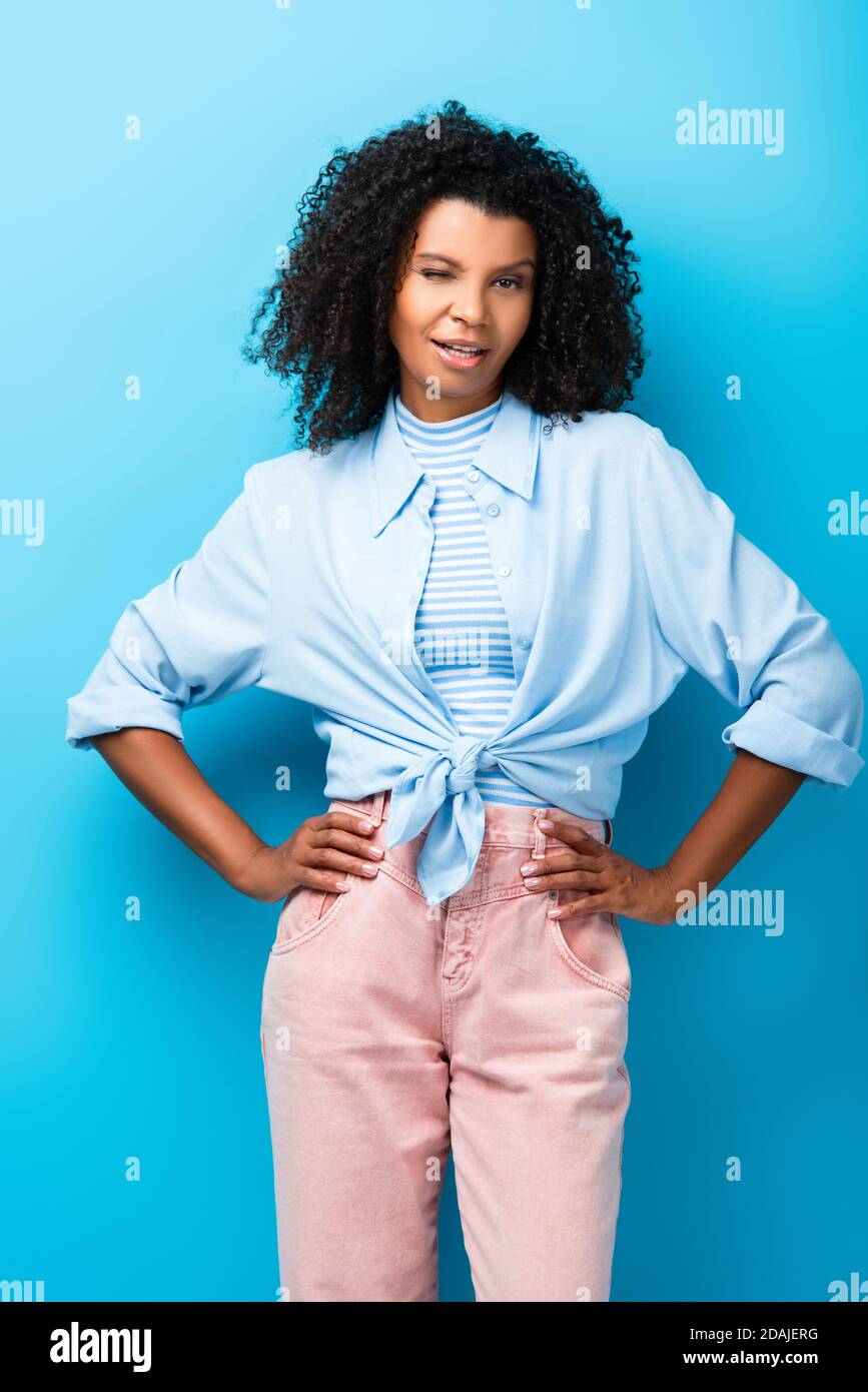 Lockige afroamerikanische Frau, die mit der Hand auf Hüften steht und Augenzwinkern auf blau Stockfoto