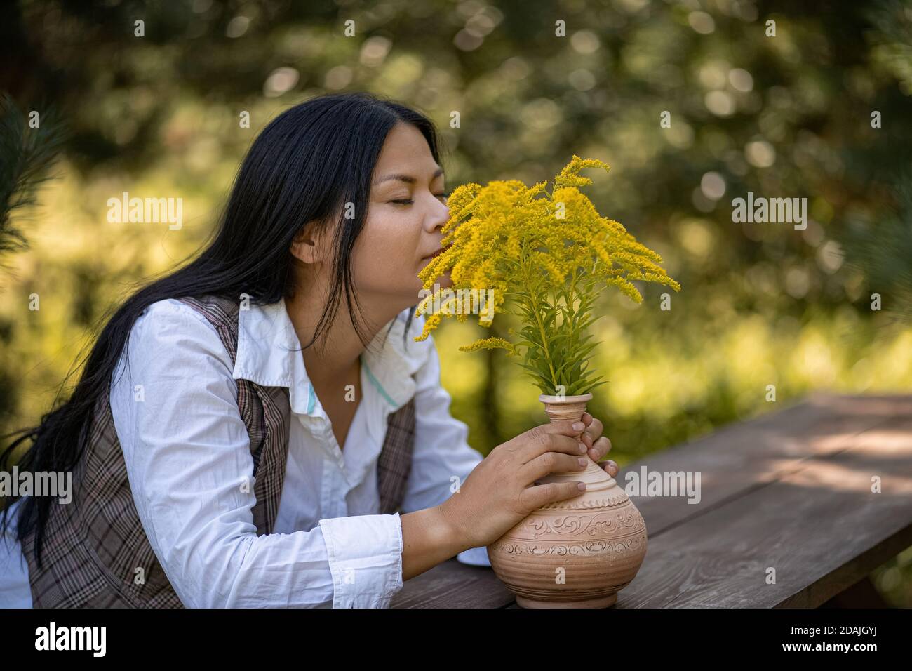 Schönheit Frau schnüffelt Duft von gelben Blumen an warmen Tag im Garten. Asiatische Brünette ruht am Holztisch mit Strauß von Solidago auf ihm. Weiblich Stockfoto