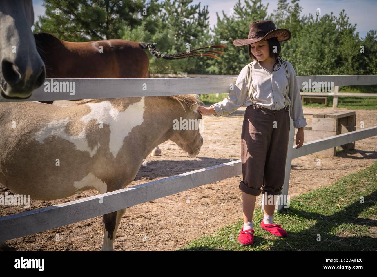 Lächelndes Mädchen in Cowboy-Kleidung streichelt kleines Pony im Paddock am sonnigen Sommertag. Betreuung der Tiere Konzept Stockfoto