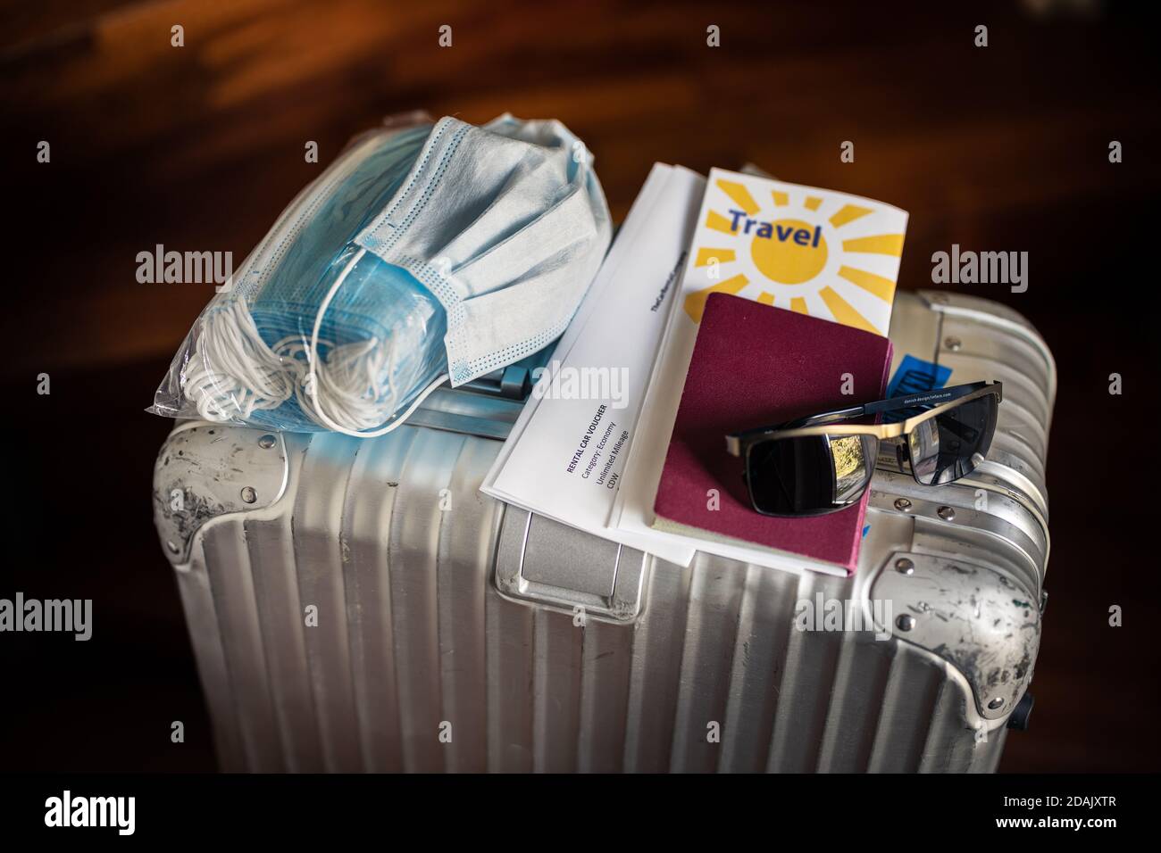 Reisen in Zeiten des Corona Virus Konzept: Ein silberner Koffer mit einer Packung Masken, eine gebrauchte Maske, Pass, Reisedokumente und Sonnenbrille auf der Oberseite. Stockfoto