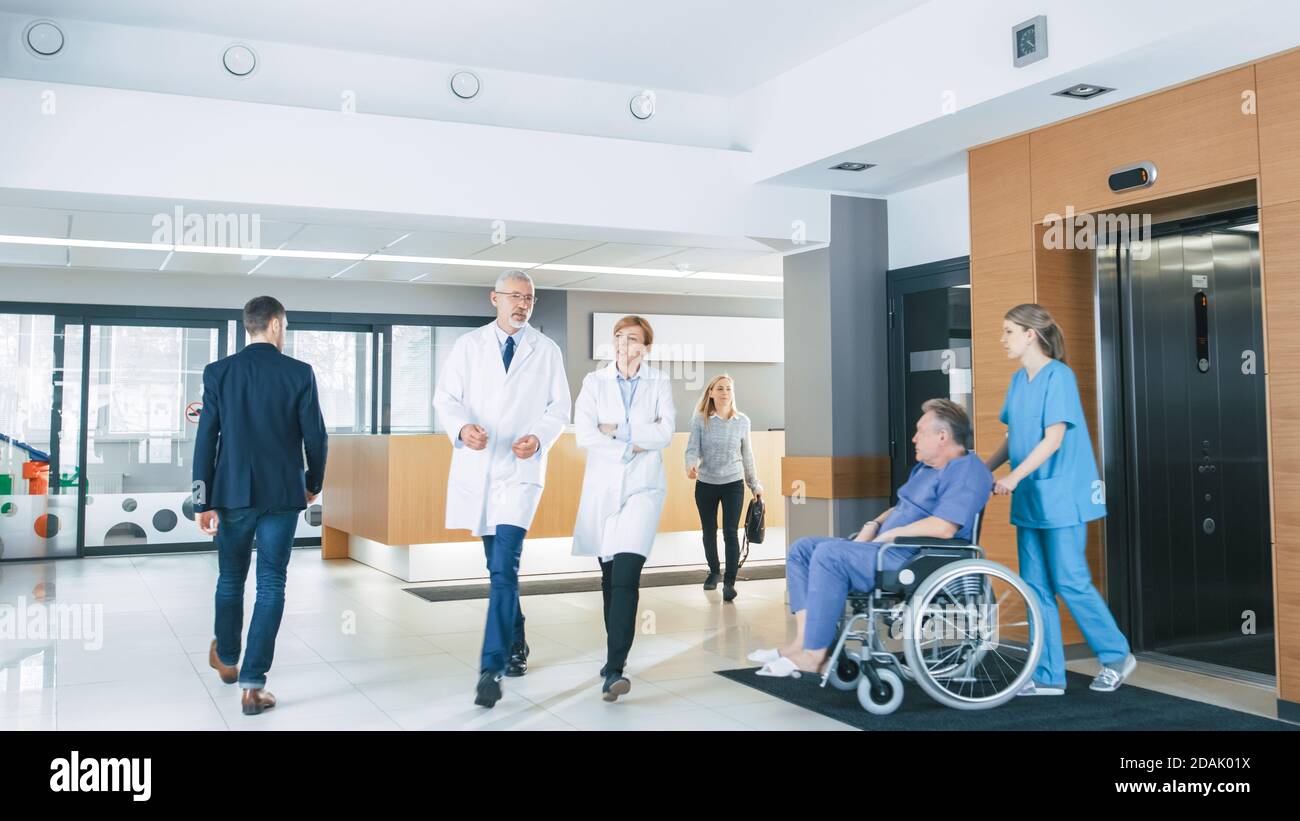 Erster Stock des geschäftigen Krankenhauses, Ärzte, Krankenschwestern und Personal beschäftigt arbeiten, Assistent bewegt älteren Mann im Rollstuhl. Neue Moderne Medizin Stockfoto
