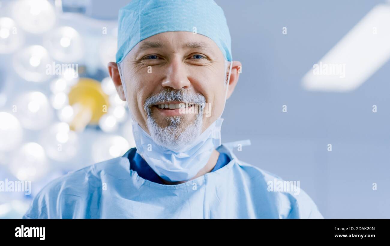 Portrait des professionellen Chirurgen hebt OP-Maske nach erfolgreicher Operation Simling in die Kamera. Im Hintergrund modernes Krankenhaus Stockfoto