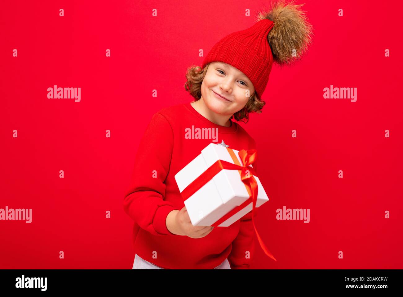 Ein cooler Junge mit Locken auf einem roten Wandhintergrund In einem Pullover mit einem Weihnachtsbaum mit einem Geschenk Box in seinen Händen Stockfoto