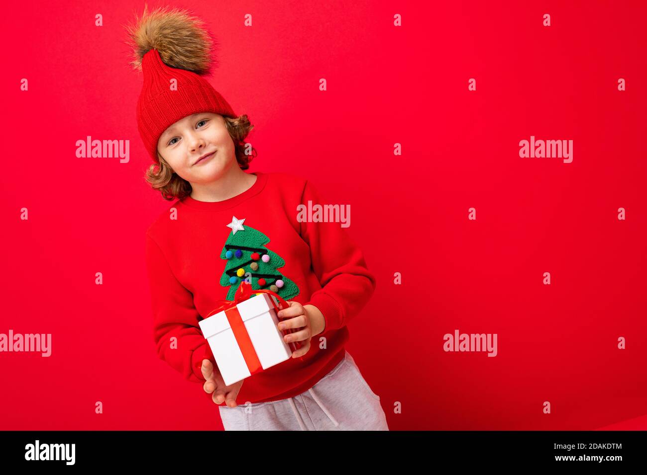 Ein cooler Junge mit Locken auf einem roten Wandhintergrund In einem Pullover mit einem Weihnachtsbaum mit einem Geschenk Box in seinen Händen Stockfoto