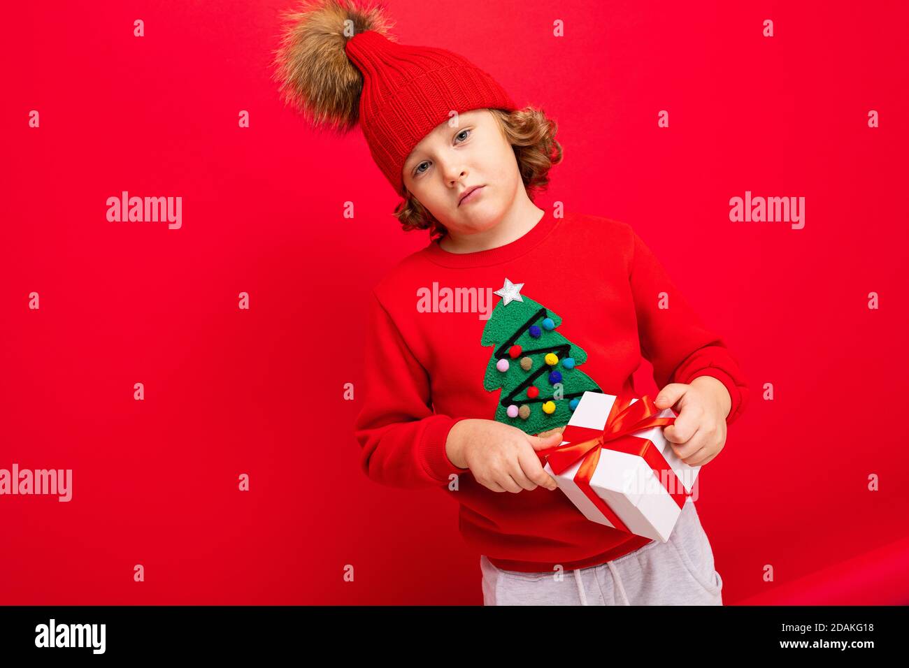 Ein cooler Junge mit Locken auf einem roten Wandhintergrund In einem Pullover mit einem Weihnachtsbaum mit einem Geschenk Box in seinen Händen Stockfoto