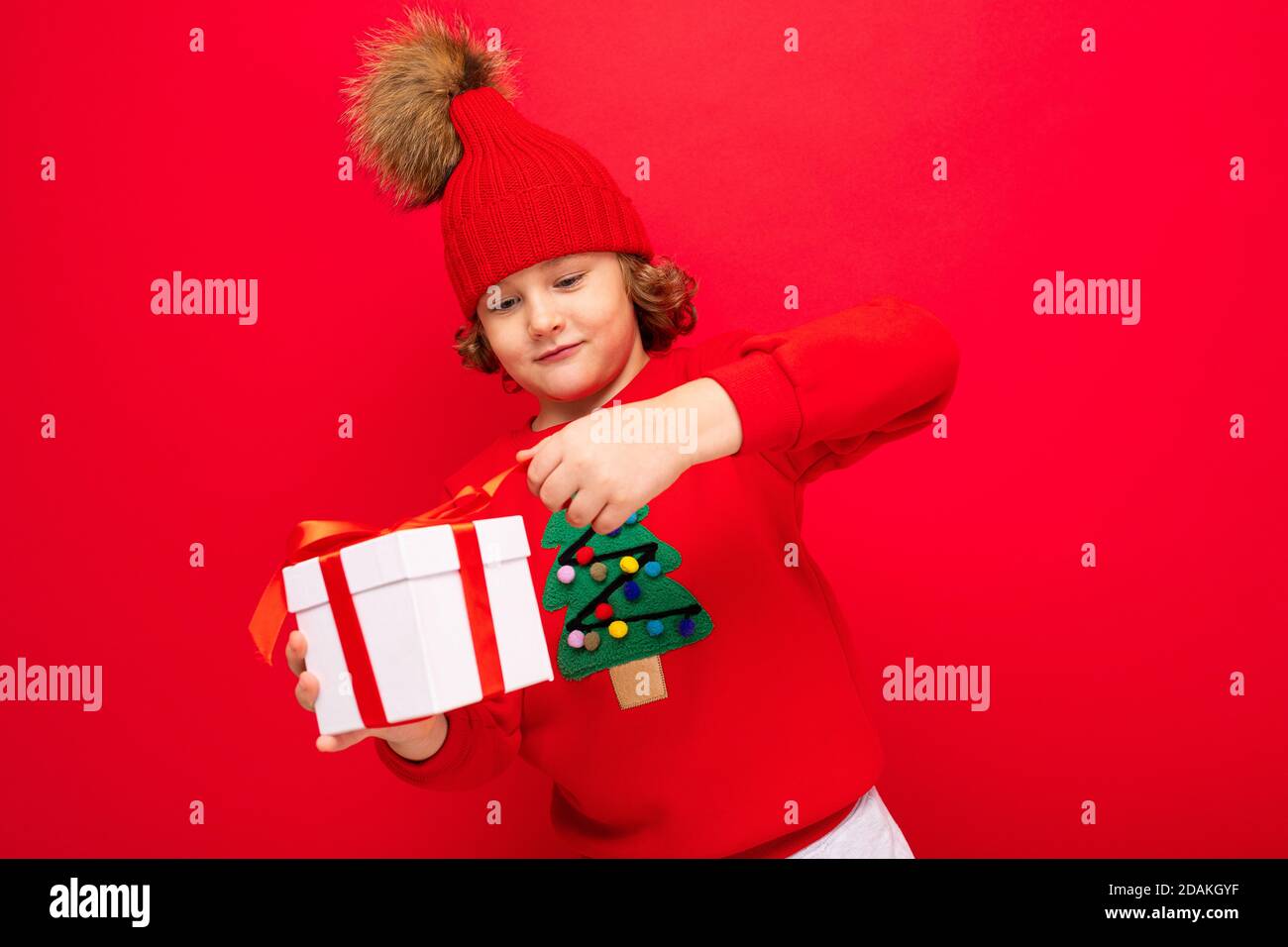 Ein cooler Junge mit Locken auf einem roten Wandhintergrund In einem Pullover mit einem Weihnachtsbaum mit einem Geschenk Box in seinen Händen Stockfoto