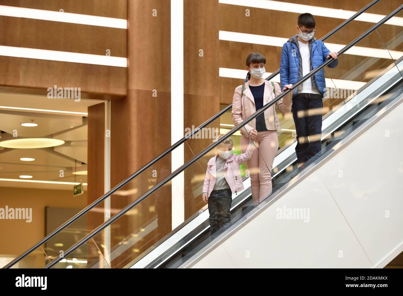 Mama und Kinder mit Gesichtsmasken fahren die Rolltreppe hinein Das Einkaufszentrum während der Quarantäne Stockfoto