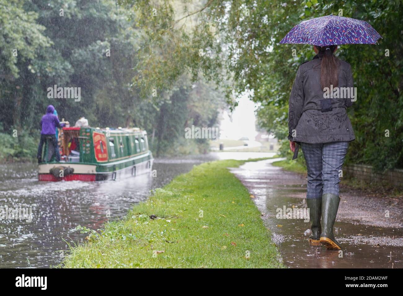 Rückansicht einer Frau mit Regenschirm, die im Regen neben einem Schmalboot auf dem britischen Kanal läuft. Stockfoto