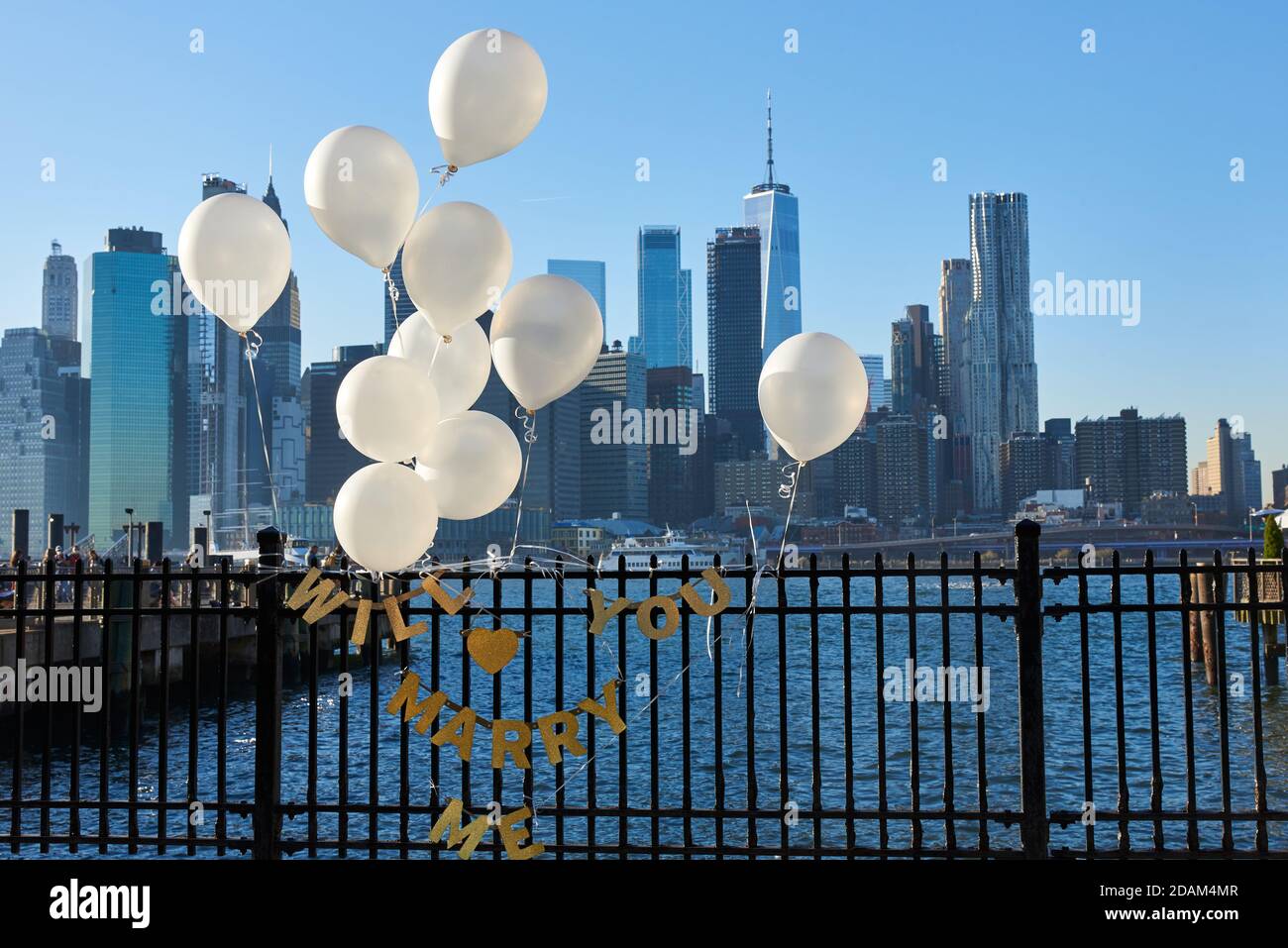 Ein Hochzeitsvorschlag mit weißen Ballons und goldenen Schriftzügen auf einem Zaun im Brooklyn Bridge Park, mit dem East River Stockfoto