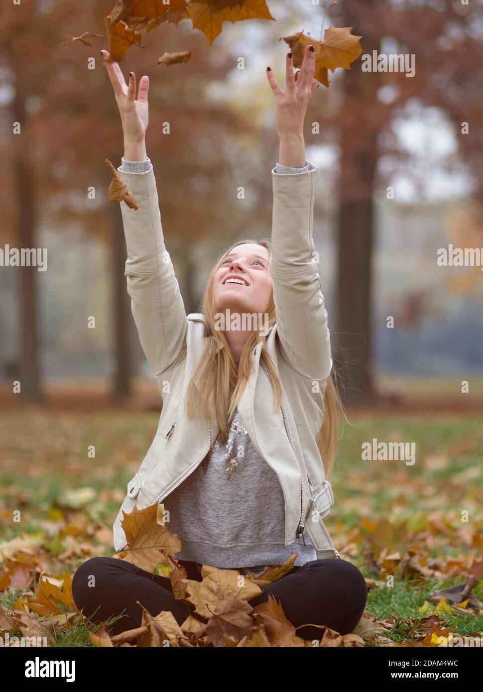 Junge Blonde Frau sitzt im Herbst Park werfen braunen Ahorn Blätter Stockfoto