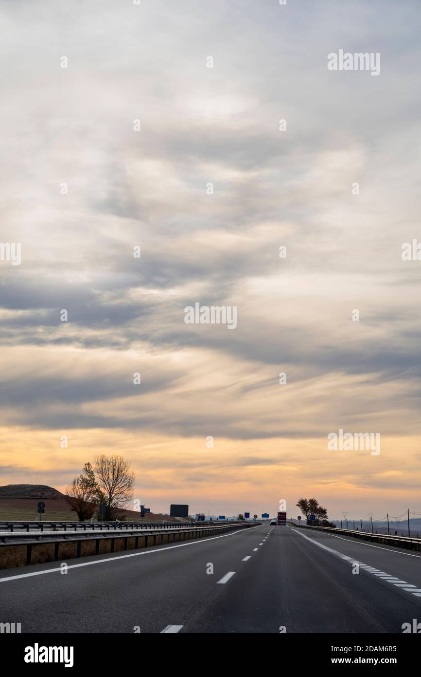 Eine Autobahngeraden, die sich gegen einen wolkigen Sonnenaufgangshimmel im vertikalen Format verirrt, überholt ein Auto einen Lastwagen weiter die Straße hinunter. Stockfoto