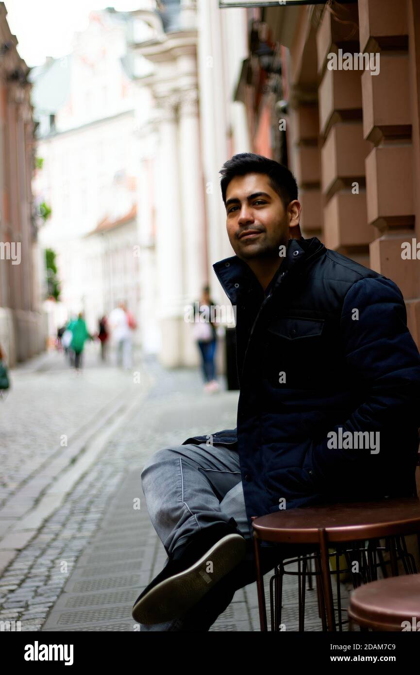 Ein südasiatischer indischer Ethnie Mann, der in einem offenen Straßenrestaurant in Mitteleuropa sitzt. Low-Angle-Aufnahme eines lächelnden Mannes, der die Kamera anschaut Stockfoto