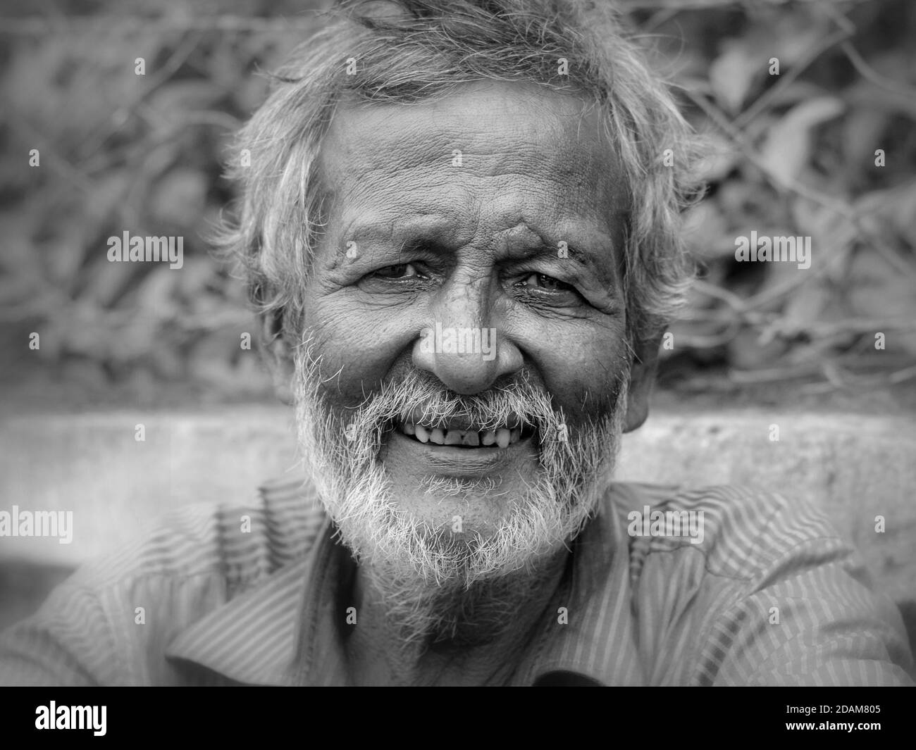 Ältere indische Wanderarbeiterin mit kurzem Bart sitzt am Straßenrand, wartet auf einen täglichen Job und lächelt für die Kamera. Stockfoto