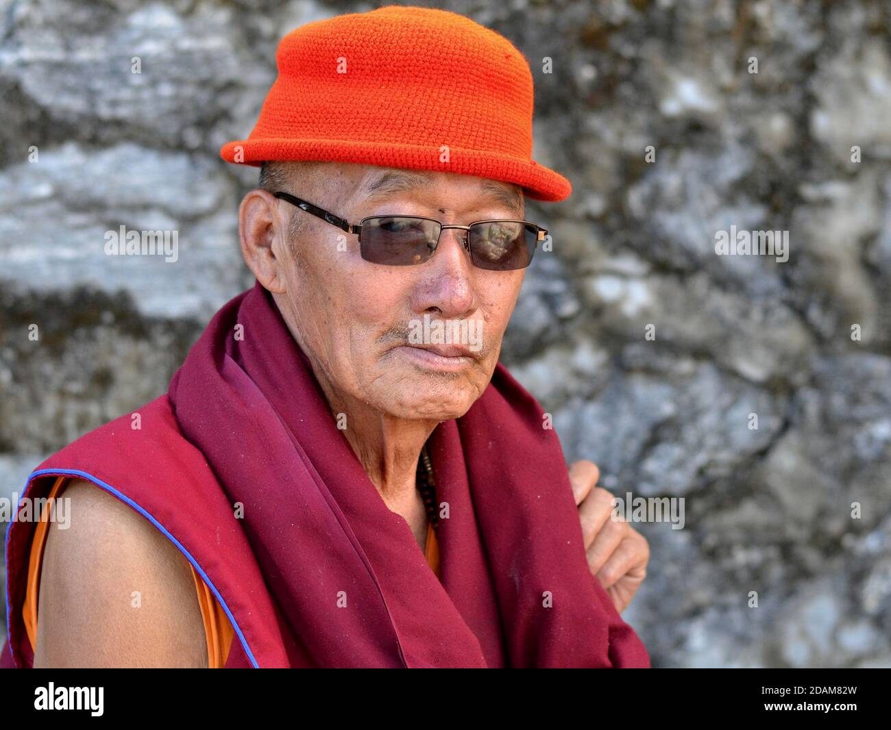 Älterer tibetischer buddhistischer Mönch mit billiger moderner Sonnenbrille trägt einen gestrickten orangefarbenen Hut und eine kastanienbraune Robe und posiert für die Kamera. Stockfoto