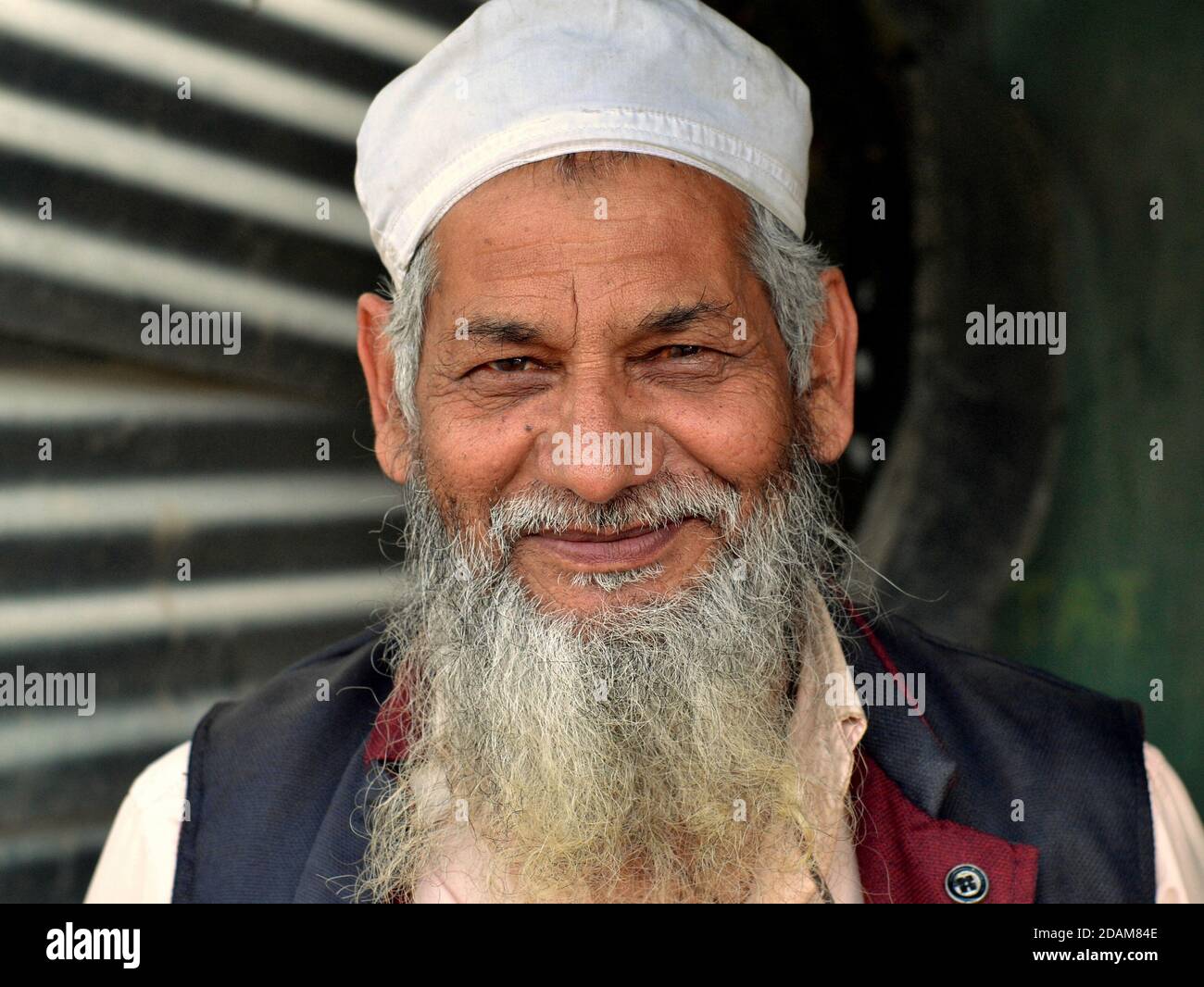 Ältere nordindische muslimische Ladenbesitzer mit islamischem Bart und weißer Gebetsmütze posiert und lächelt für die Kamera. Stockfoto