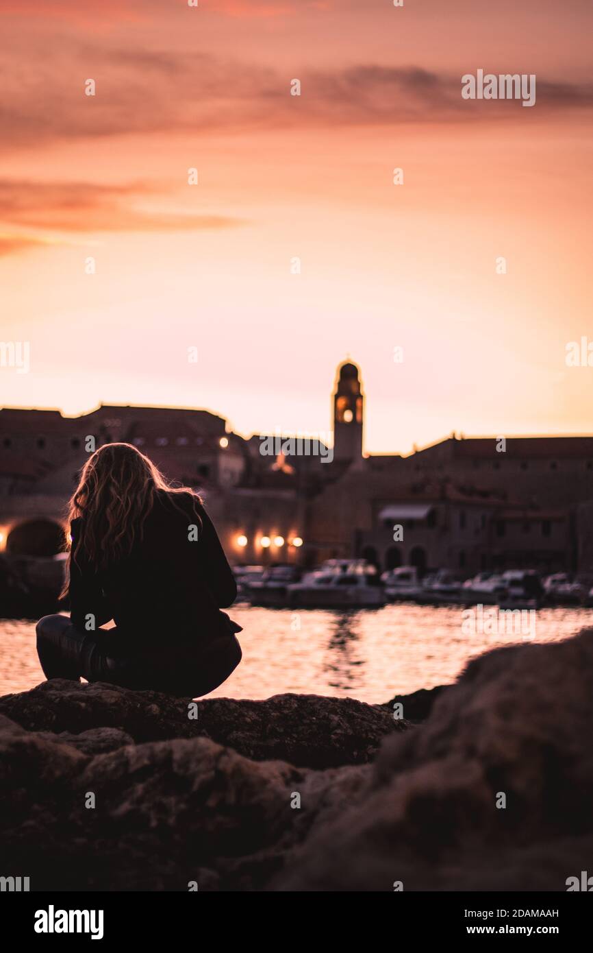 Schöner Sonnenuntergang über der Altstadt von Dubrovnik. Kleiner lokaler Hafen vor der Stadt, Frau in schwarzen Kleidern sitzt auf einem nahe gelegenen Felsen und beobachtet t Stockfoto