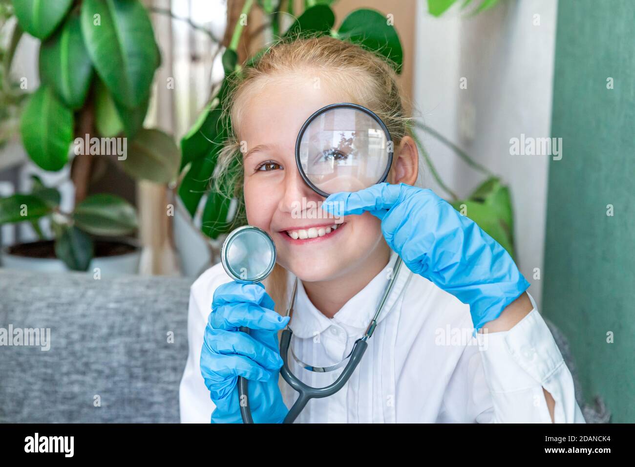 Ein kaukasisches Mädchen von 9-10 Jahren in einem weißen Mantel, blaue medizinische Handschuhe hält ein Stethoskop in der Hand, schaut durch eine Lupe, lächelt. Res Stockfoto