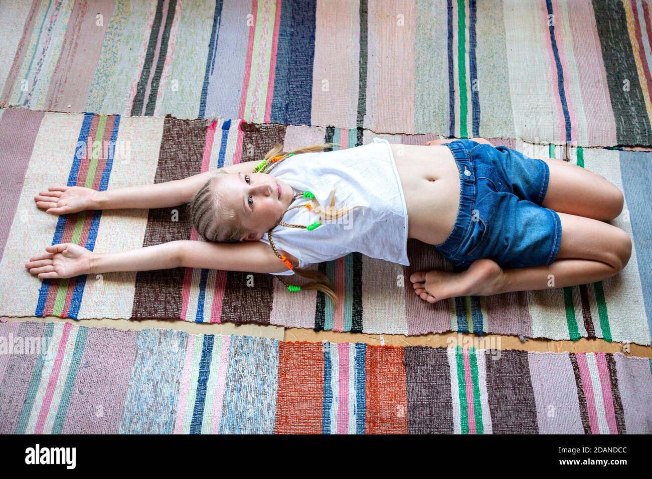 Kaukasische Mädchen mit Zöpfen in einem weißen T-Shirt macht Yoga, Stretching zu Hause auf einem gestreiften Stoff Teppich.Digital Detox, Wiedervereinigung mit der Natur, gesund br Stockfoto