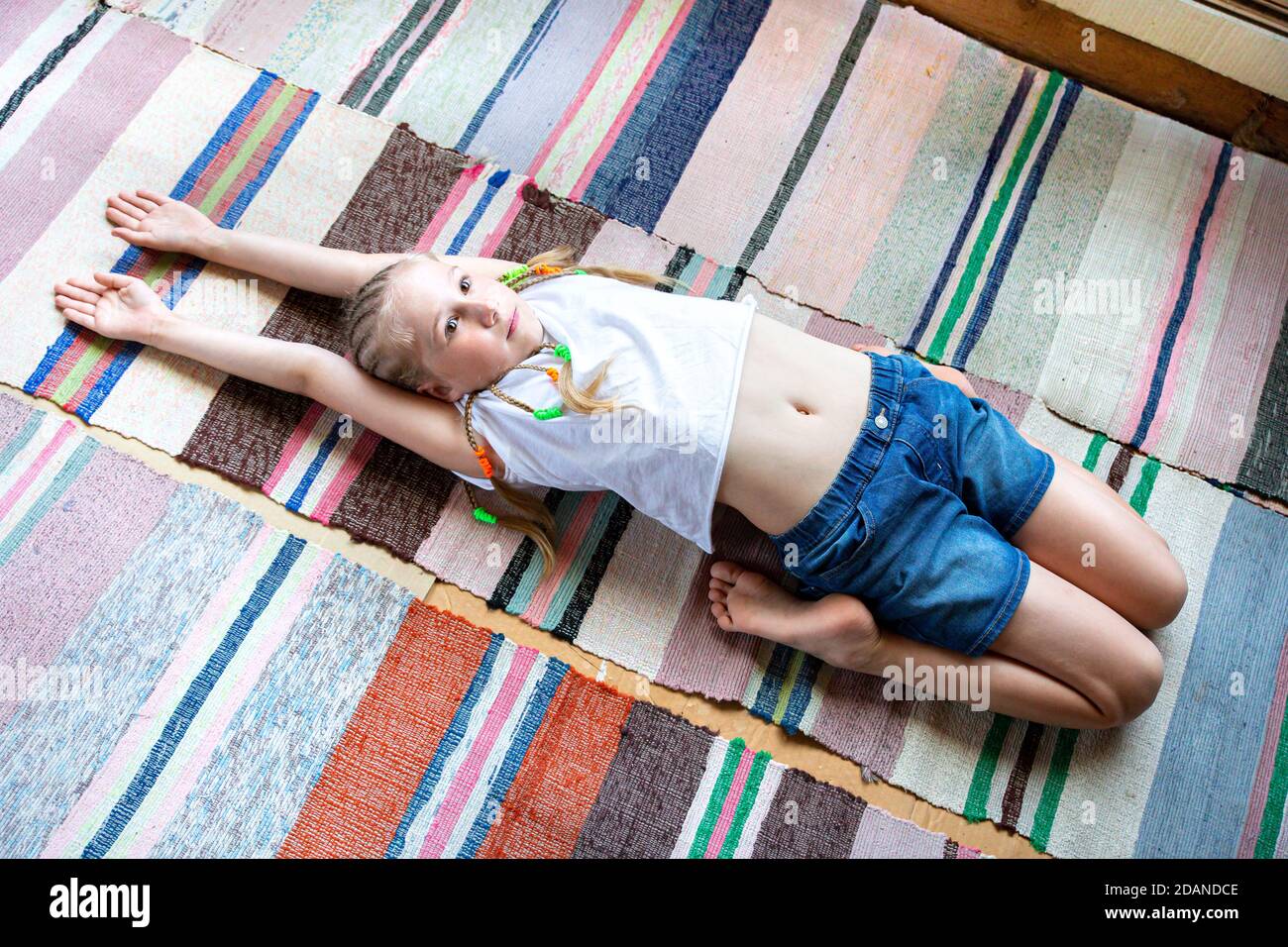 Kaukasische Mädchen mit Zöpfen in einem weißen T-Shirt macht Yoga, Stretching zu Hause auf einem gestreiften Stoff Teppich.Digital Detox, Wiedervereinigung mit der Natur, gesund br Stockfoto