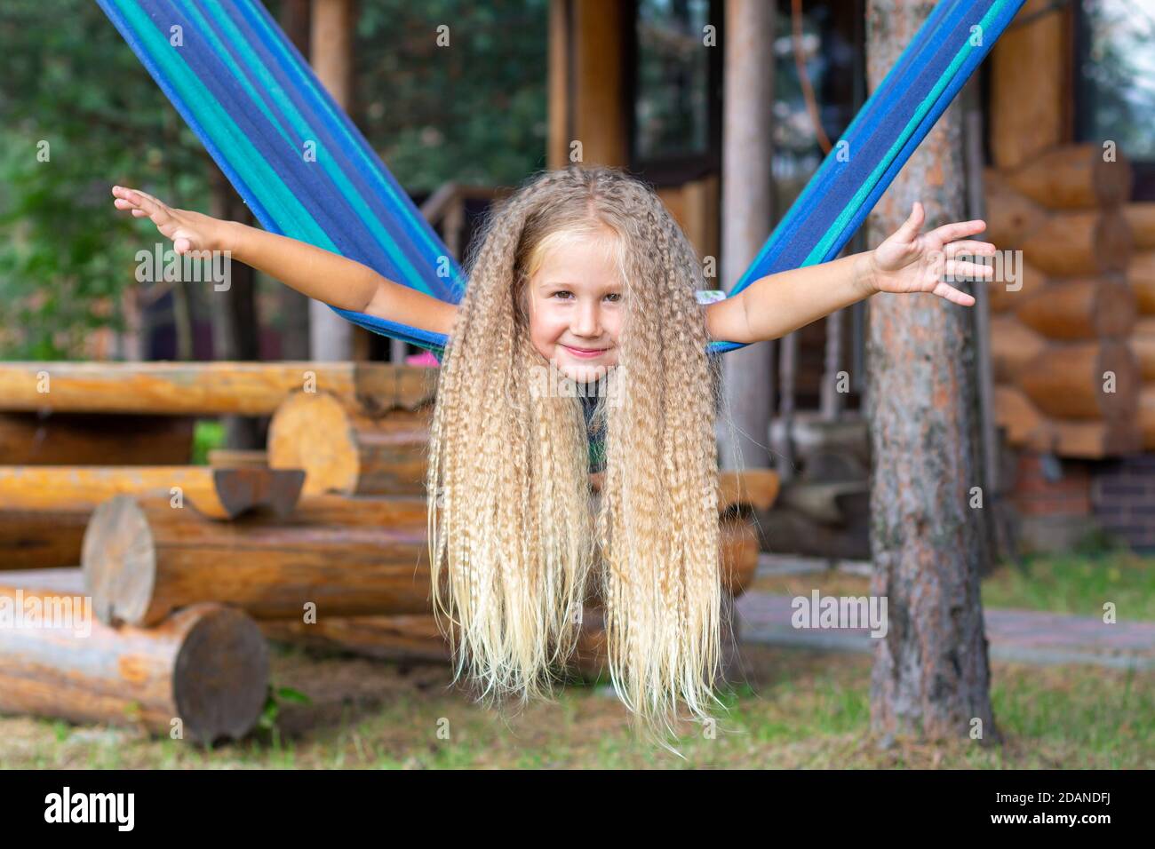 Kleines glückliches Mädchen mit langen blonden lockigen Haaren schwingt die Arme auf einer blau-grünen Hängematte ausgestreckt. Bewegungsfreiheit, Lebensstil. Schulferien, Urlaub Stockfoto