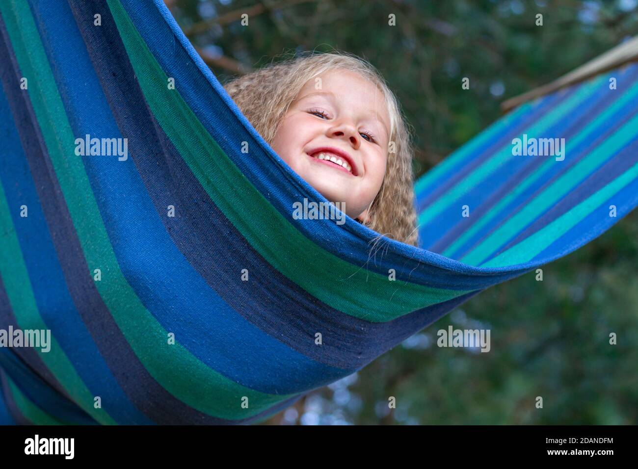 Kleines glückliches kaukasisches Mädchen mit blondem lockiges Haar schwankt auf mehrfarbig gestreifter Hängematte. Porträt eines Kindes, Nahaufnahme. Peekaboo. Lifestyle. Sorglos Stockfoto