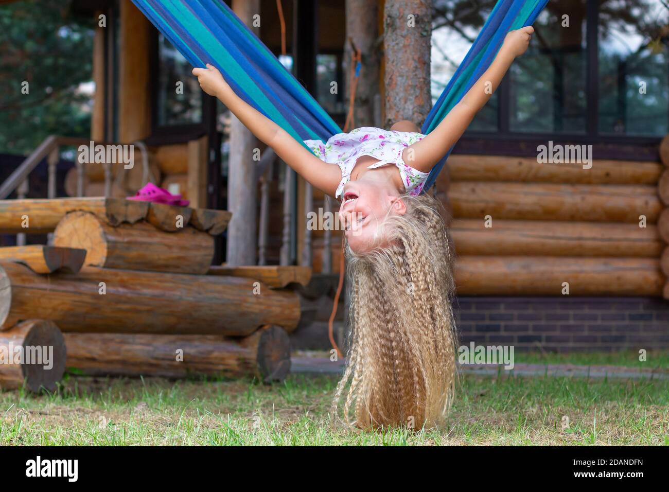 Kleines glückliches Mädchen mit langen blonden lockigen Haaren schwänzt kopfüber auf einer blau-grünen Hängematte. Bewegungsfreiheit, Lebensstil. Schulferien, Urlaub, Tag Stockfoto
