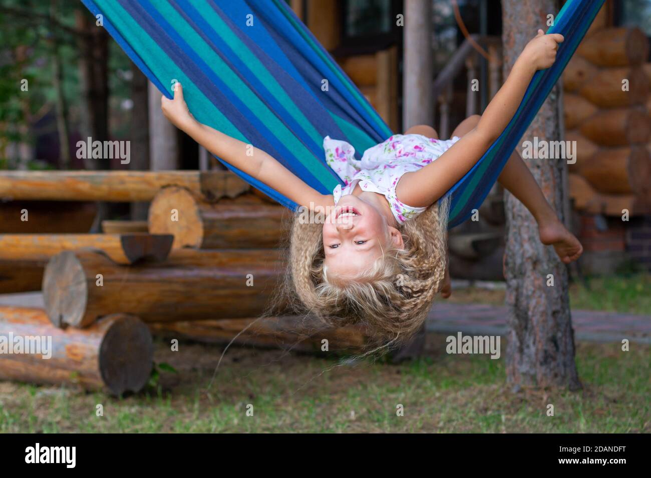Kleines glückliches Mädchen mit langen blonden lockigen Haaren schwänzt kopfüber auf einer blau-grünen Hängematte. Bewegungsfreiheit, Lebensstil. Schulferien, Urlaub, Tag Stockfoto