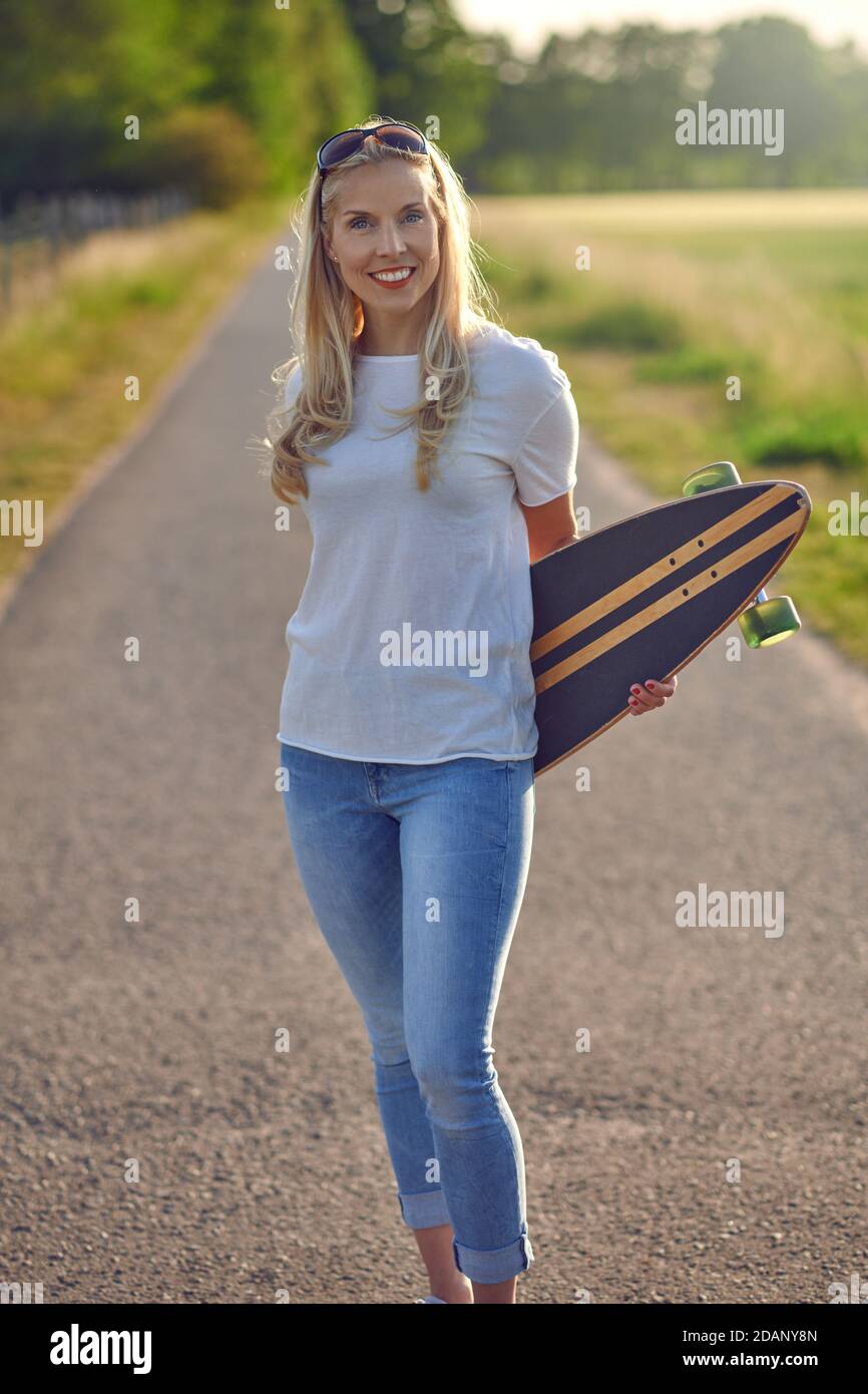 Porträt einer fit schöne Frau mittleren Alters mit einem aktiven Lebensstil lächeln und Blick auf die Kamera, während ein Longboard auf einer sonnigen Straße in der Stockfoto