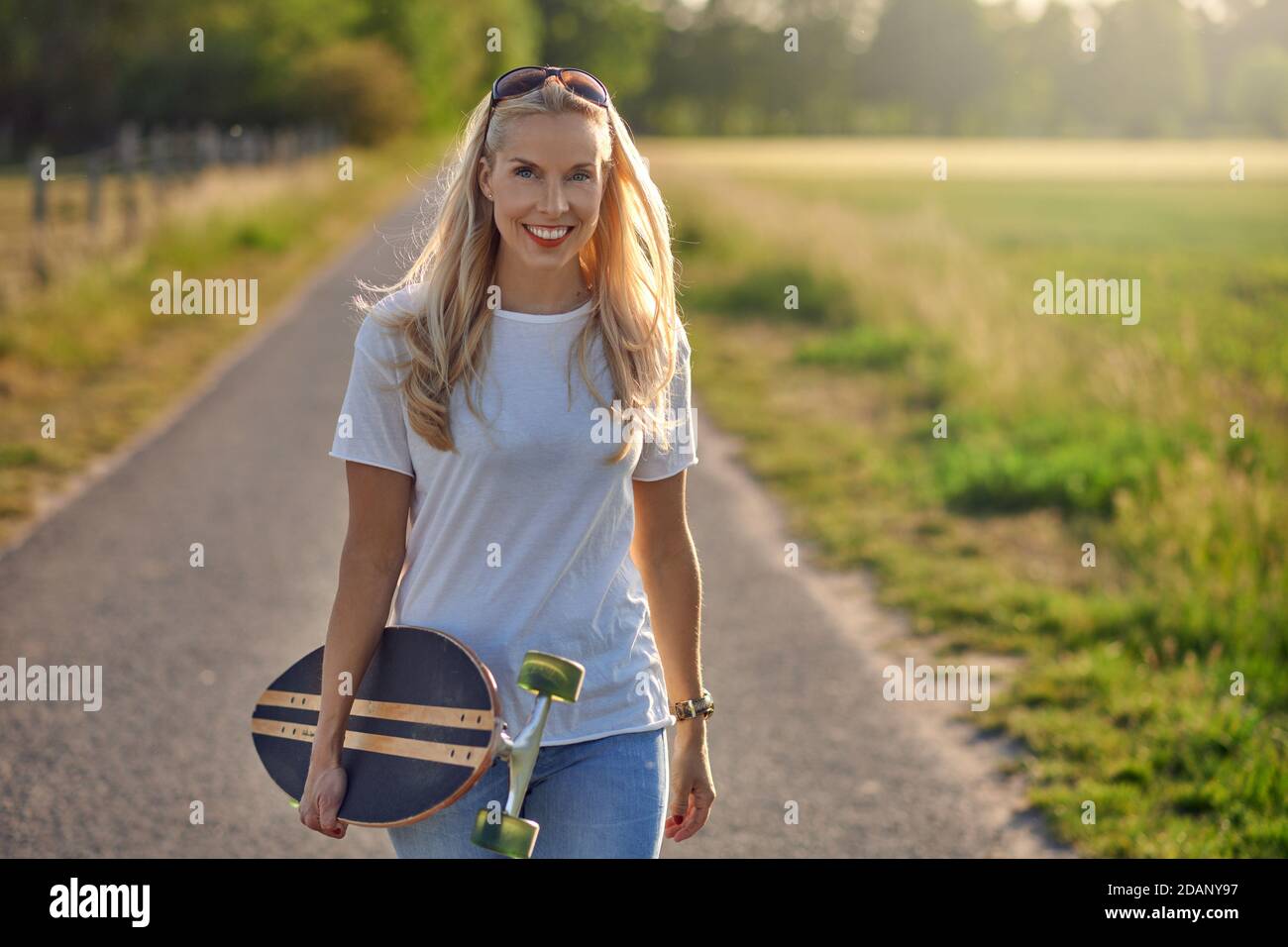 Porträt einer fit schöne Frau mittleren Alters mit einem aktiven Lebensstil lächeln und Blick auf die Kamera, während ein Longboard auf einer sonnigen Straße in der Stockfoto